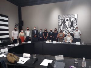 Foto colorida de reunião com o chefe de gabinete da Secretaria de Governo, Amauri Gavião. Nela temos 5 mulheres e 6 homens, tendo 10 pessoas em pé e uma em cadeira de rodas. Eles estão em volta de uma mesa com papéis e canetas. Ao fundo, parede cinza com quadro do Abaporu, do Romero Britto.